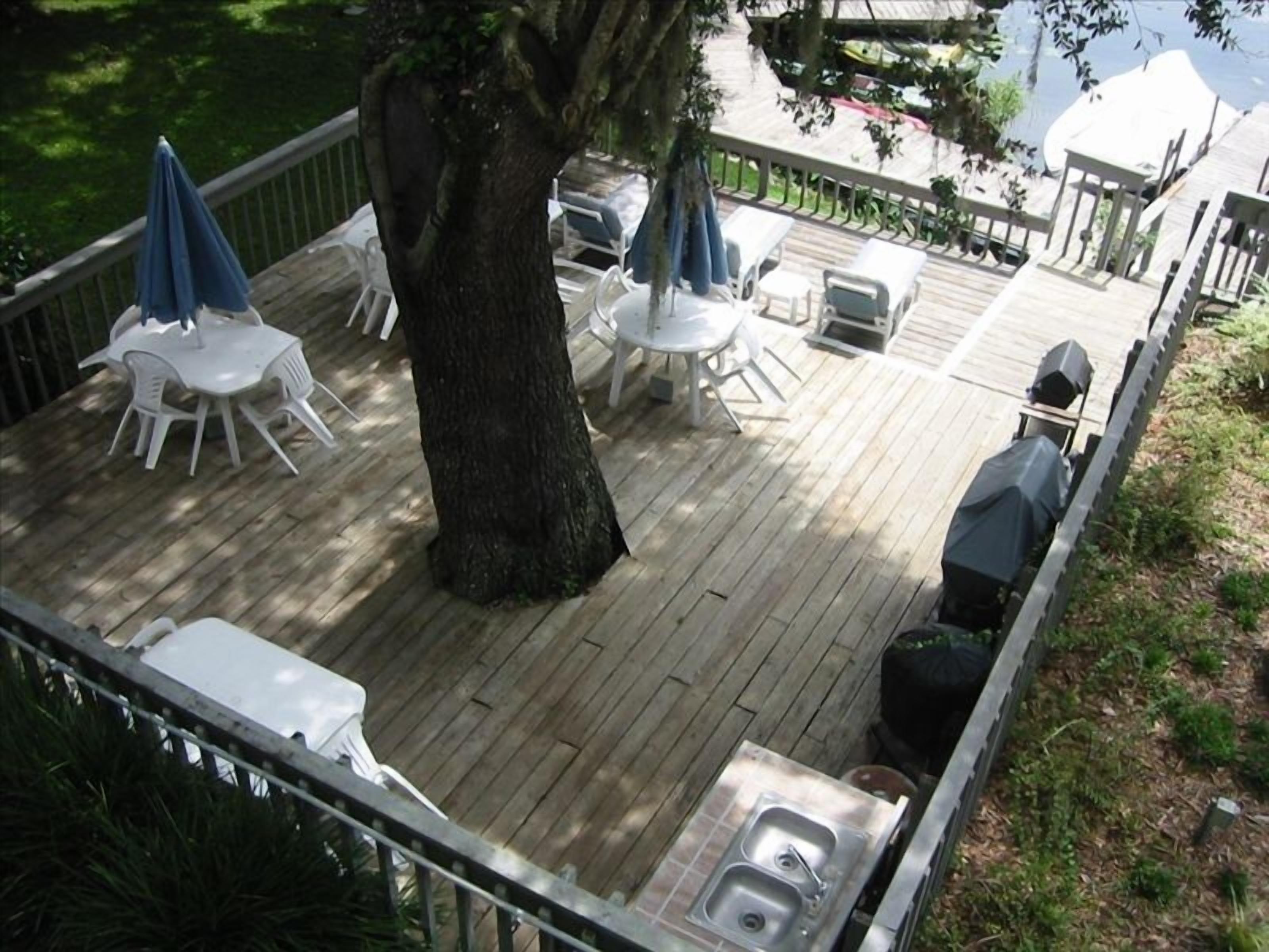 Una terraza de madera con mobiliario exterior, incluyendo sillas y mesas de plástico blanco, un lavamanos de metal y una parrilla.