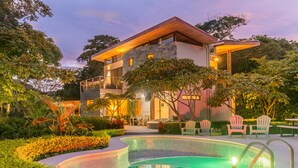 Una casa moderna de dos pisos con piscina, rodeada de exuberante vegetación y mobiliario de exterior.