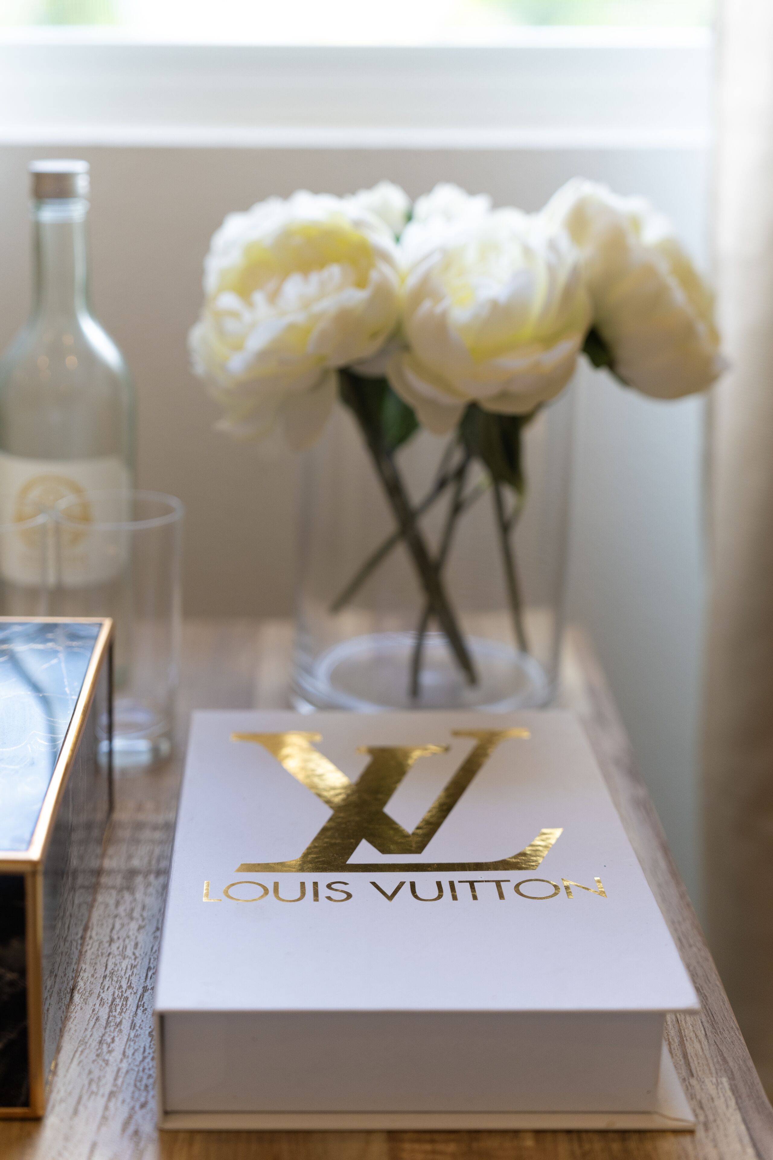 A Louis Vuitton box on a wooden table with a vase of white flowers and a clear bottle in the background.