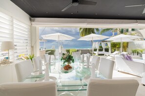 Un espace repas avec une table en verre, des chaises blanches et des parasols, donnant sur une plage.