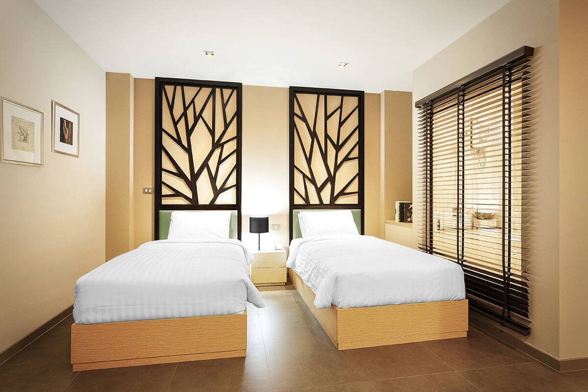 A hotel room with two beds, a large window with blinds, a decorative screen, and framed pictures on the wall.