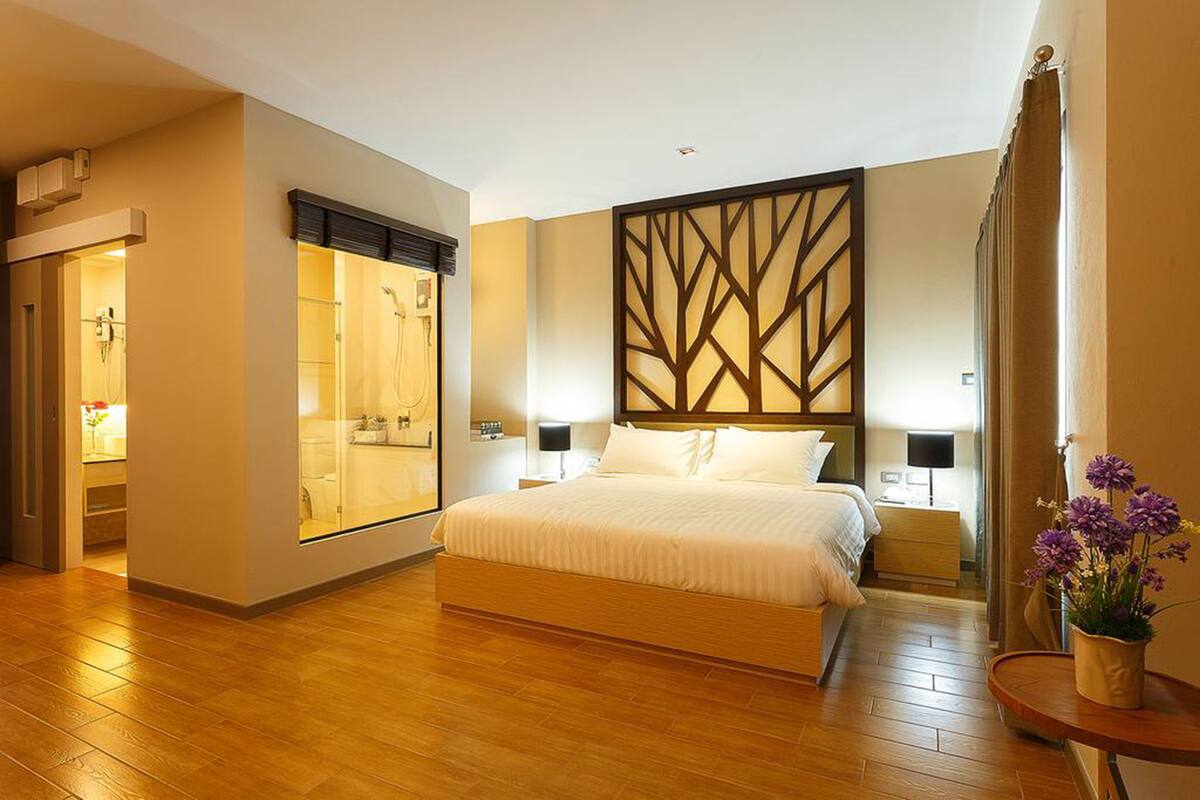 A hotel room with a large bed, a wooden floor, a bathroom with a glass shower enclosure, and a decorative wall panel.