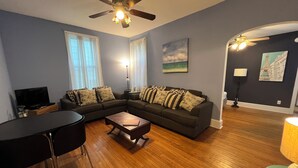 A living room with a sectional sofa, a coffee table, a TV, and a ceiling fan. - Cozy Downtown Condo(Dog Friendly) 3.5 Blocks to Beach.1 block to Washington Mall (Cape May)