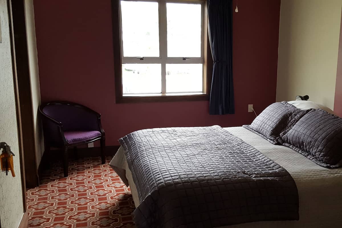 Un dormitorio con cama, una silla, un ventanal y alfombra estampada.