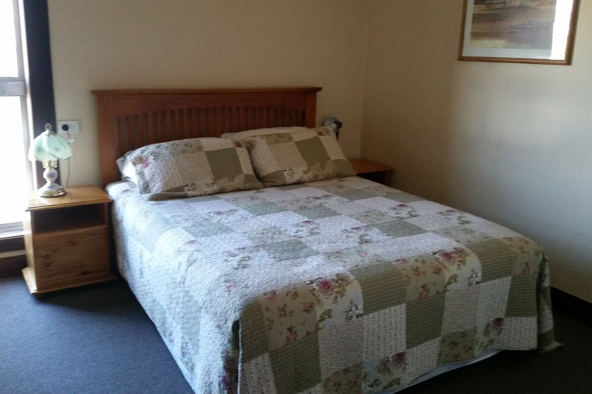 Una cama bien hecha con una colcha estampada, un cabecero de madera y una lámpara de noche en una mesita.