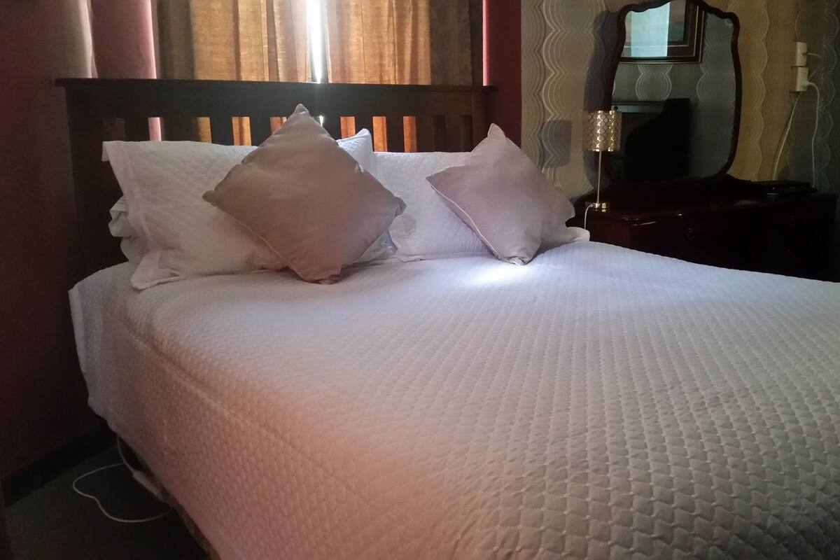 Una cama bien hecha con ropa blanca y dos almohadas, un cabecero de madera, una mesita de noche con una lámpara y un espejo en la pared.