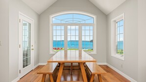 A dining area with a large table and benches, a view of the ocean through a large arched window, and a door with glass panels. - Waterfront Beach House - 2700 Sq Ft, Fiber, mooring (Friday Harbor)