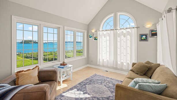 A spacious living room with a large window offering a view of the sea and a grassy landscape. There is a brown sofa, a white side table, and a patterned rug. - Waterfront Beach House - 2700 Sq Ft, Fiber, mooring (Friday Harbor)