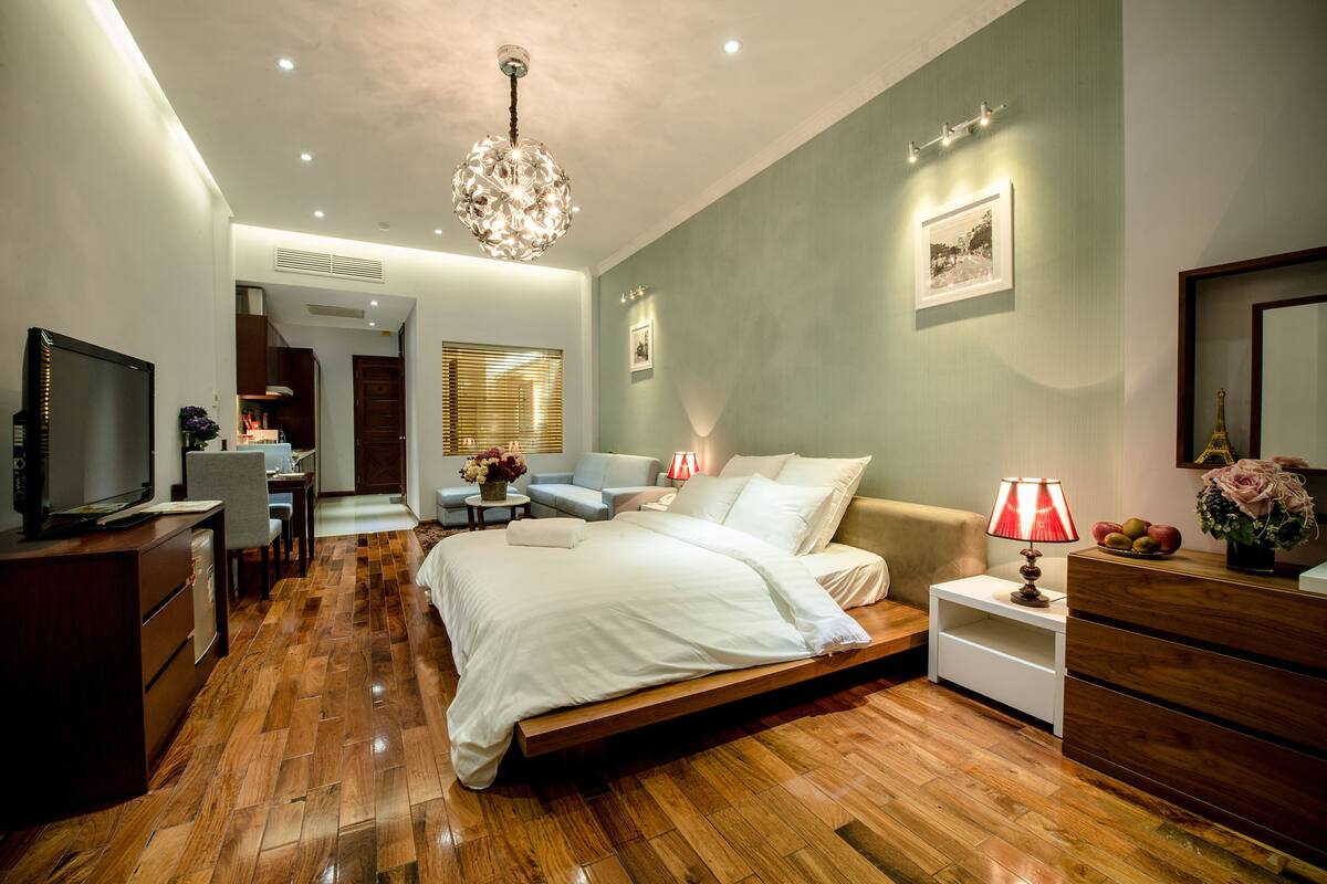 Un dormitorio moderno con piso de madera, una cama con ropa blanca, un escritorio con computadora, un televisor y una araña.