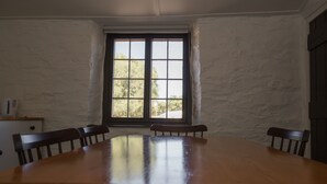 A dining room with a large wooden table and four chairs, a window with a view of trees, and a white brick wall. - Paxton Square Cottages (Burra)