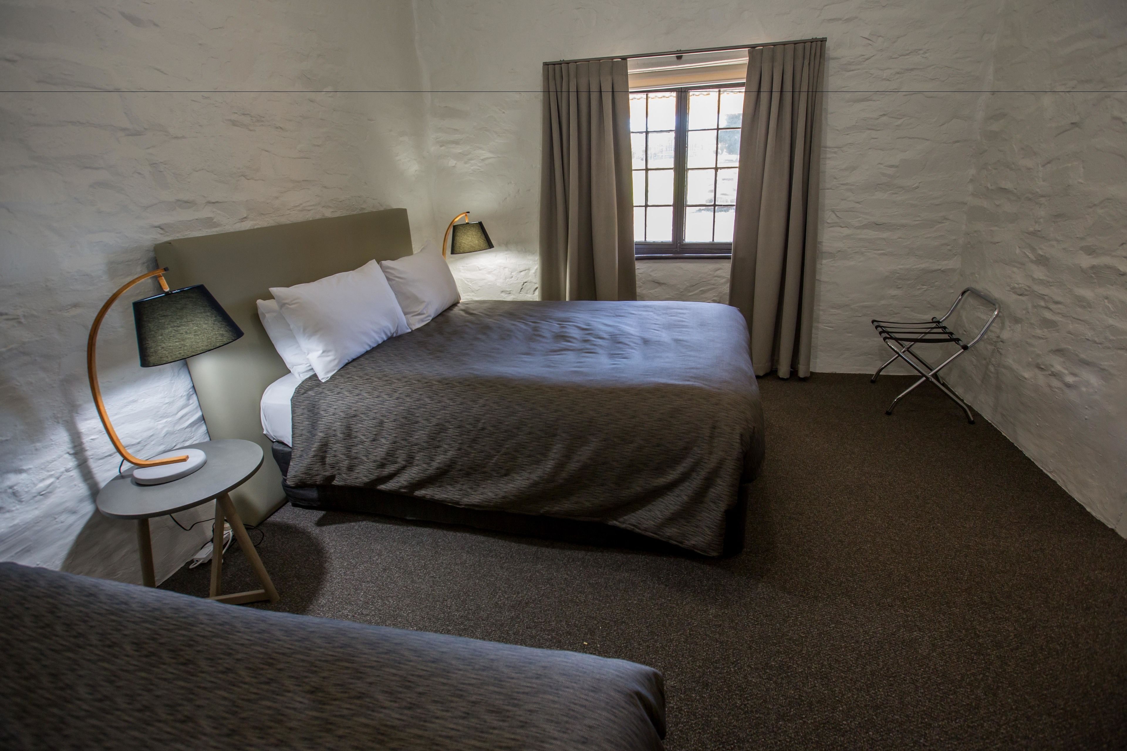 A hotel room with two beds, a small table with a lamp, a chair, and a window with curtains.
