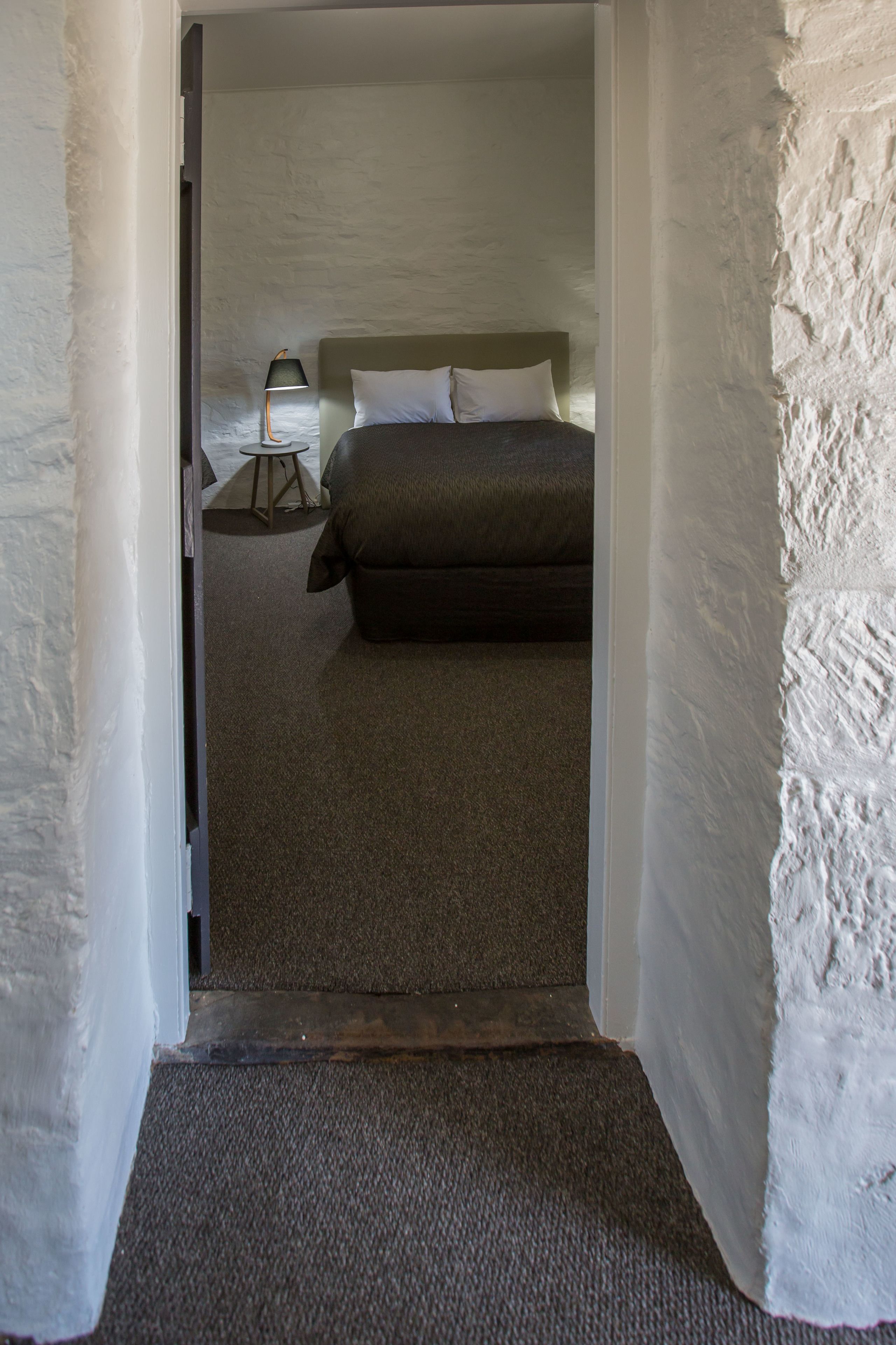 A narrow hallway leading to a bedroom with a bed, a nightstand, and a lamp.