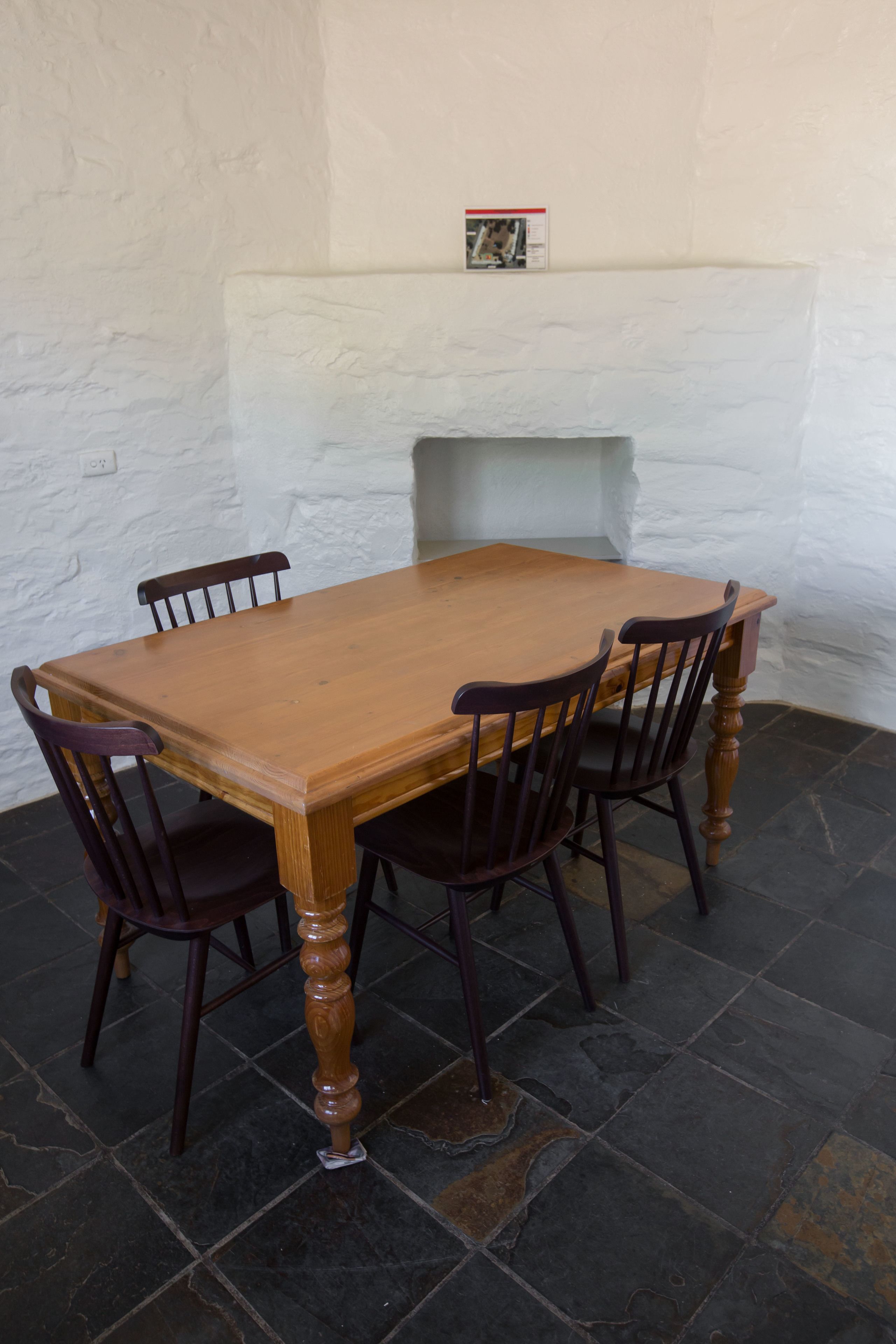Une table rectangulaire en bois, accompagnée de quatre chaises, est placée contre un mur blanc à texture, avec un petit tableau encadré au-dessus.