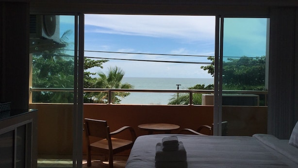 A hotel room with a bed, a chair, a small table, and a balcony with a view of the sea and palm trees. - Marina Jomtien Beach (Pattaya)