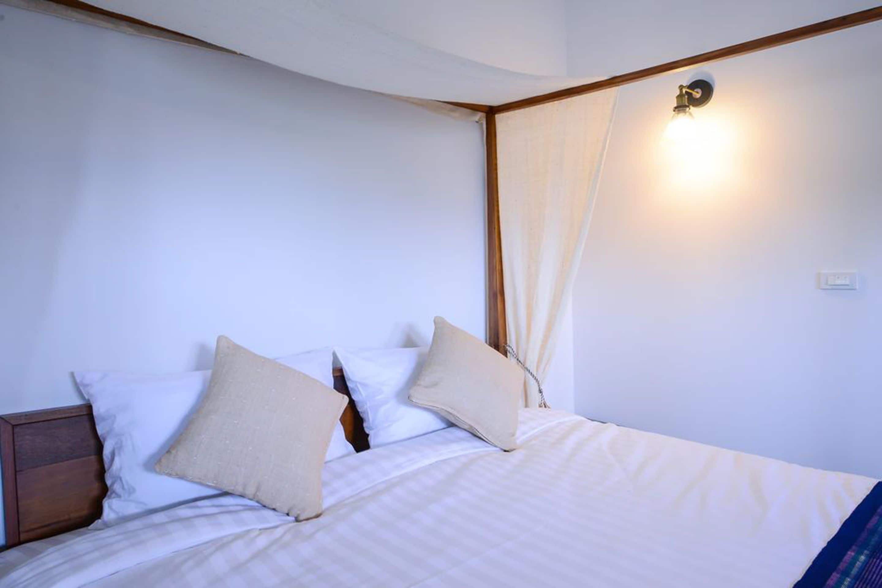 A neatly made bed with white bedding and beige pillows, a wooden headboard, and a wall-mounted light fixture.