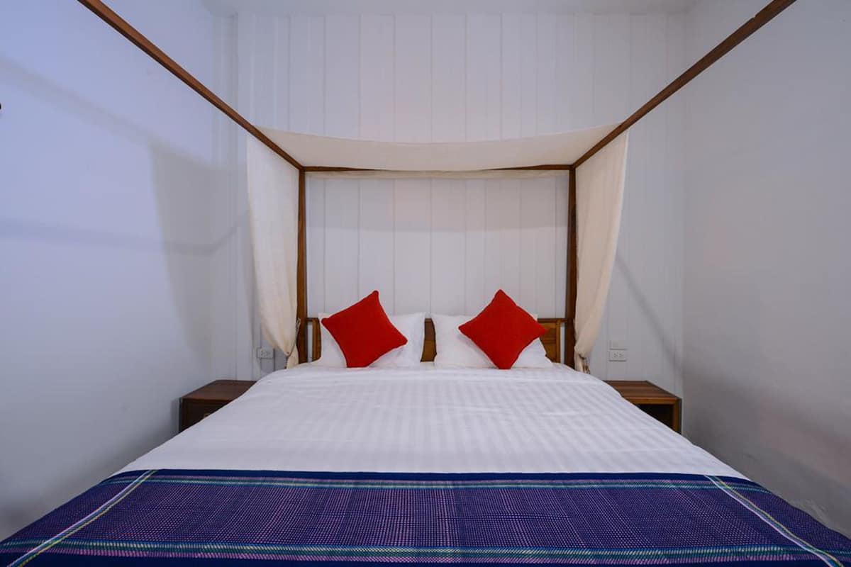 A neatly made bed with a white duvet and two red pillows, a blue patterned bedspread, and a wooden nightstand with a lamp.