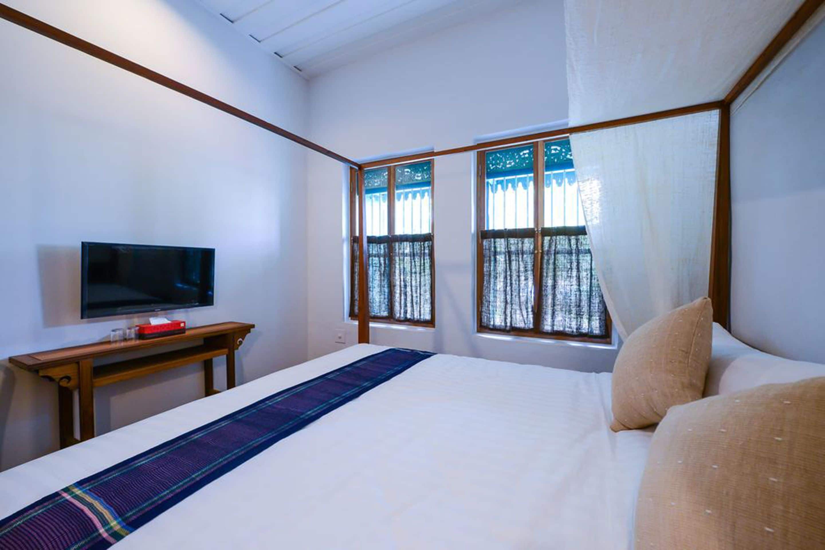Una habitación de hotel con una cama, un televisor sobre una mesita de madera y dos ventanas con cortinas.