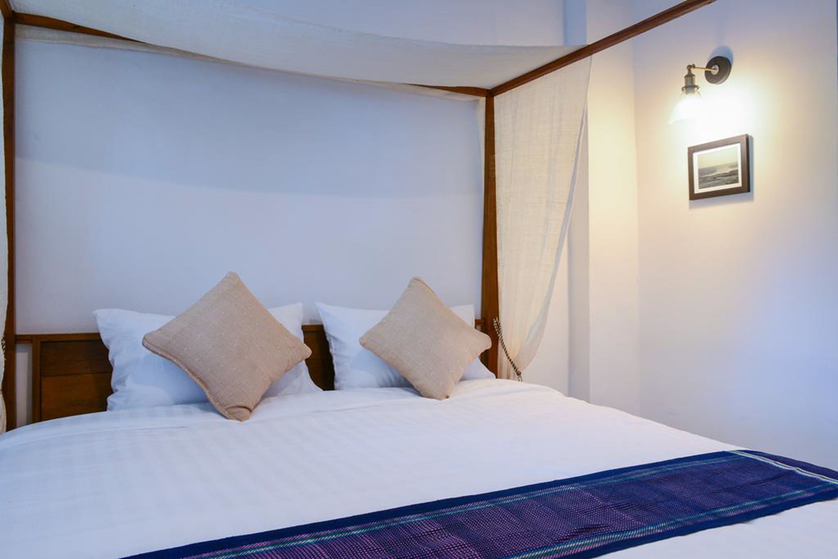 Una cama bien hecha con sábanas blancas y dos almohadas color beige, un plaid estampado en azul, una lámpara fijada a la pared y un cuadro enmarcado.
