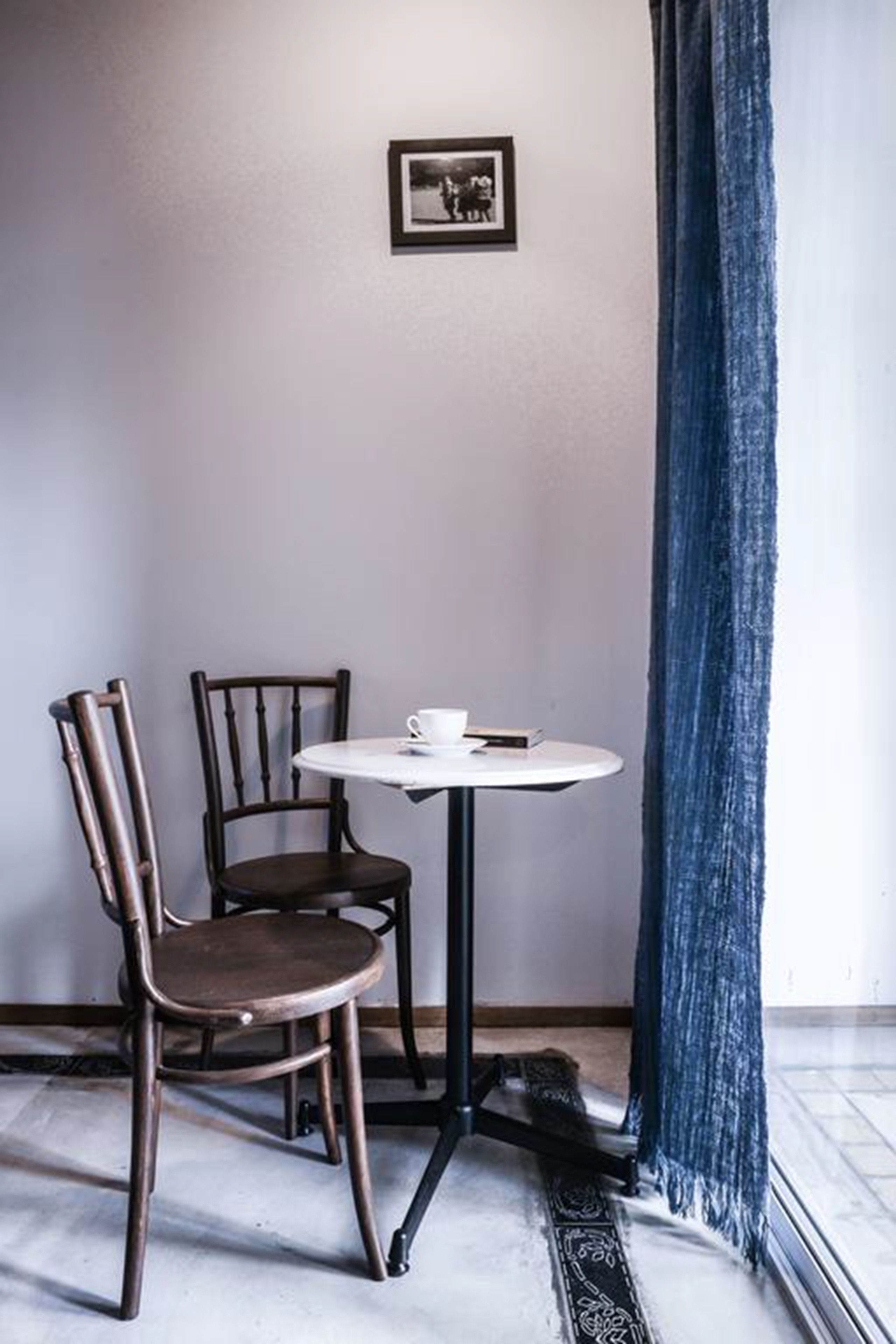 A small room with a round table, two chairs, a framed picture on the wall, and a blue curtain.
