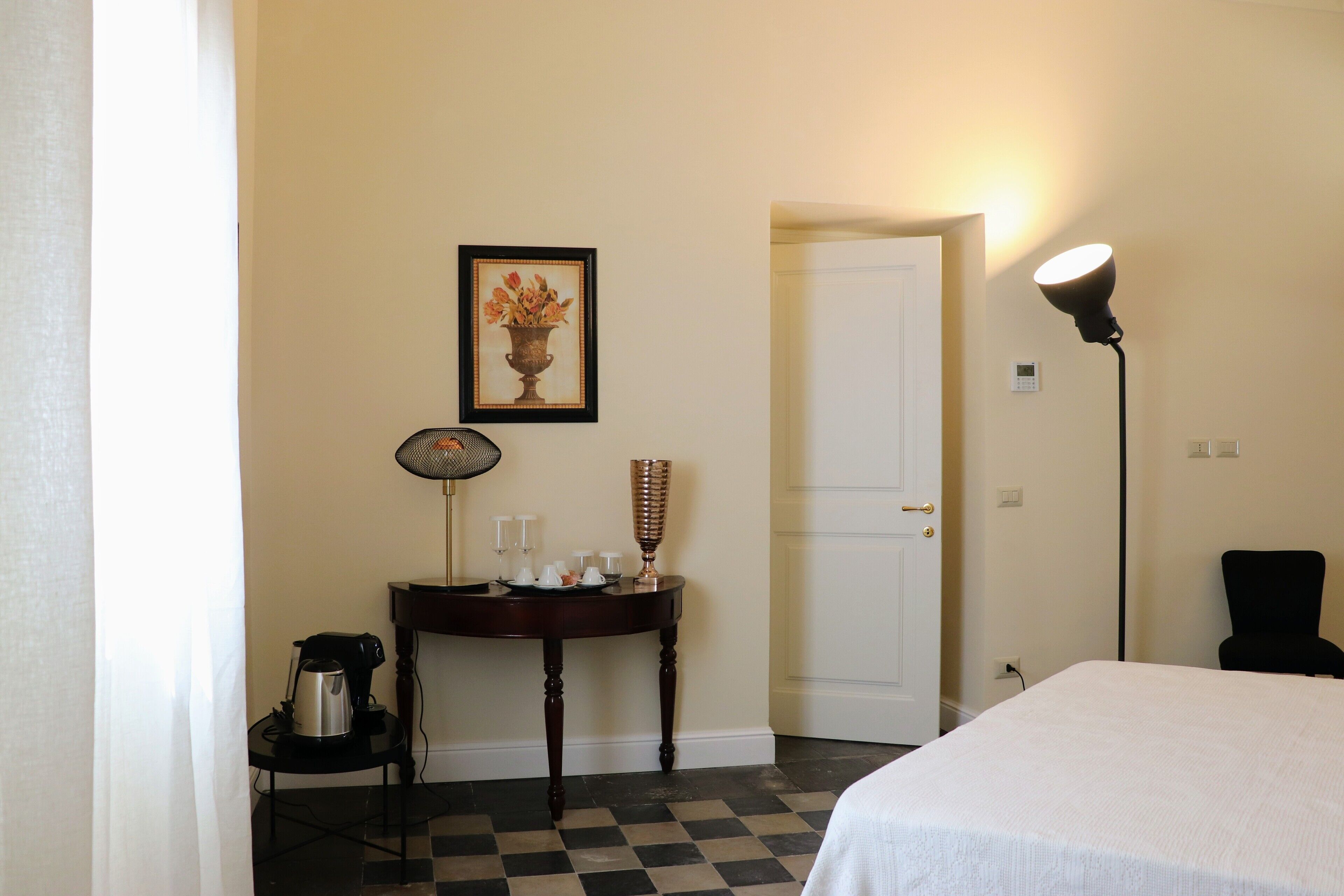 Une chambre d’hôtel avec un sol à carreaux, un lit, une table avec une lampe, un tableau au mur et une porte.