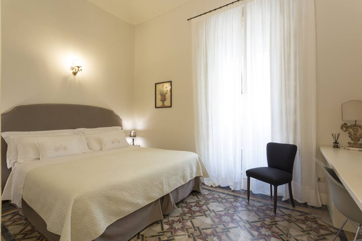 Un lit bien fait, avec une tête de lit, une chaise, un bureau et une fenêtre avec des rideaux.