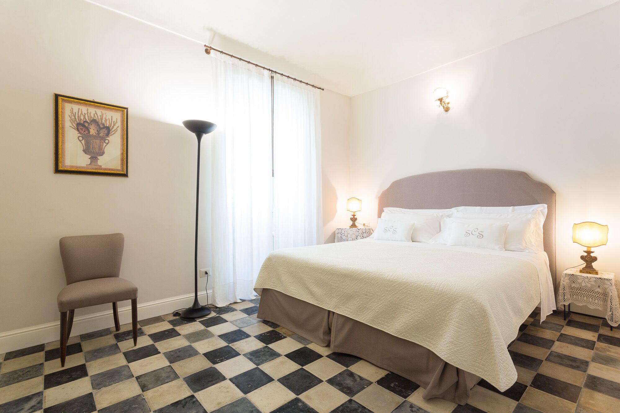 Una habitación de hotel con piso a cuadros, una cama con sábanas blancas, dos lámparas de noche, una silla y un cuadro en la pared.