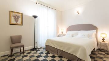 Una habitación de hotel con piso a cuadros, una cama con sábanas blancas, dos lámparas de noche, una silla y un cuadro en la pared.