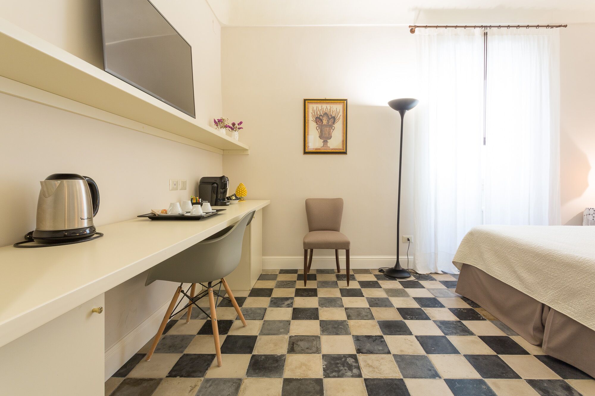 A hotel room with a checkered floor, a bed, a desk with a kettle and cups, a chair, a picture on the wall, and a floor lamp.