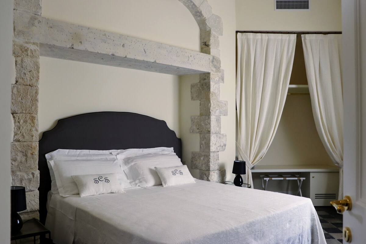 Un lit avec du linge de lit blanc et une tête de lit sombre, une arche en pierre et une fenêtre avec des rideaux blancs.