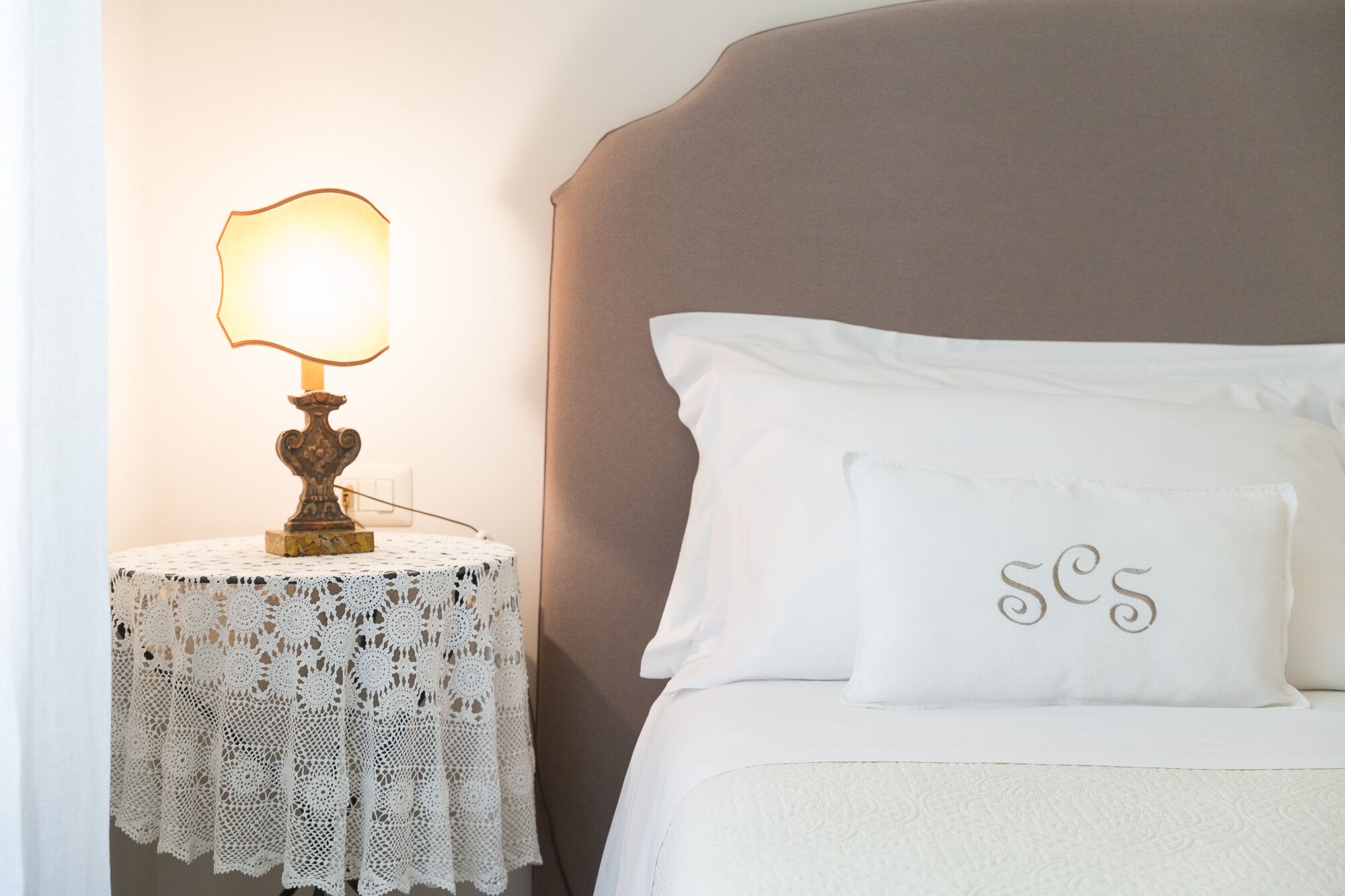 A neatly made bed with a white pillow featuring a monogram, a bedside table with a lace tablecloth, and a table lamp with a warm light.