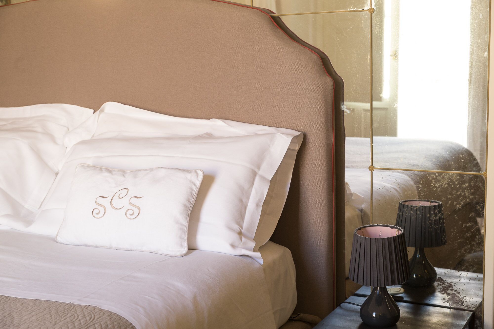A neatly made bed with white linens and a pillow with embroidered initials. A dark lamp and a small table with a vase are visible beside the bed.