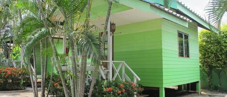 A green house with a porch, surrounded by tropical plants and flowers.