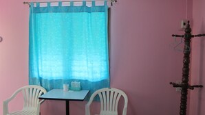 A room with two white plastic chairs, a small table, a window with a blue curtain, and a wooden stand with a hook. - The Green Resort Sri Racha (Si Racha)