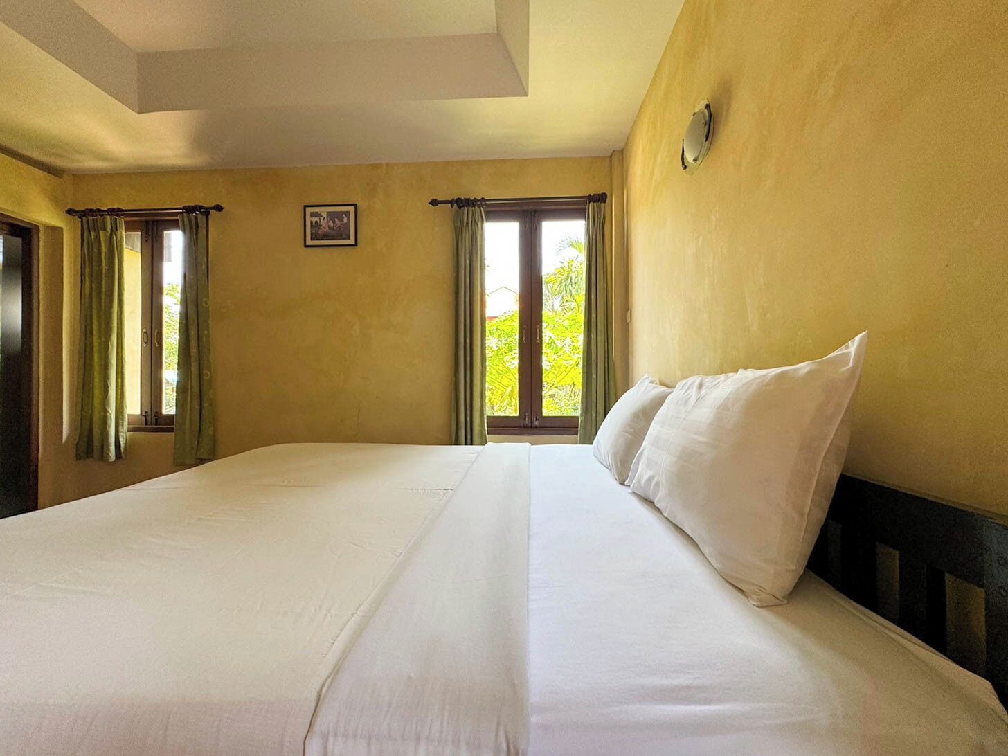 Un letto grande con lenzuola bianche in una stanza dalle pareti gialle e due finestre con tende.