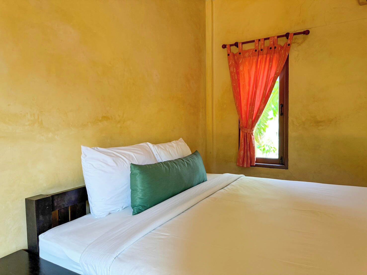 Una cama con almohadillas blancas y verdes, un cabecero de madera y una ventana con cortinas anaranjadas.