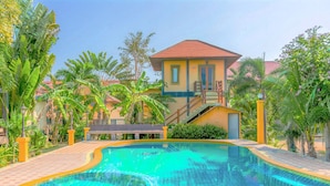 A swimming pool with a curved edge, surrounded by palm trees and a building with a red roof. - Baanbali Beach Resort (Pranburi)