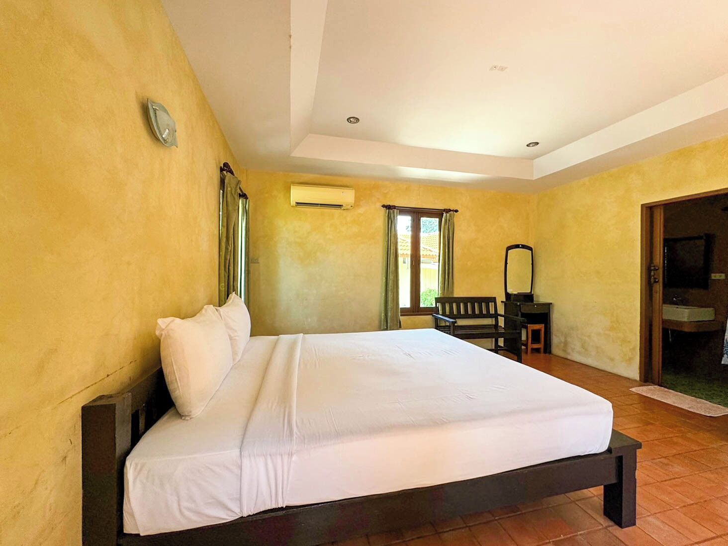 A bedroom with a large bed, a wooden bench, a small table, a mirror, and a bathroom visible through an open door.