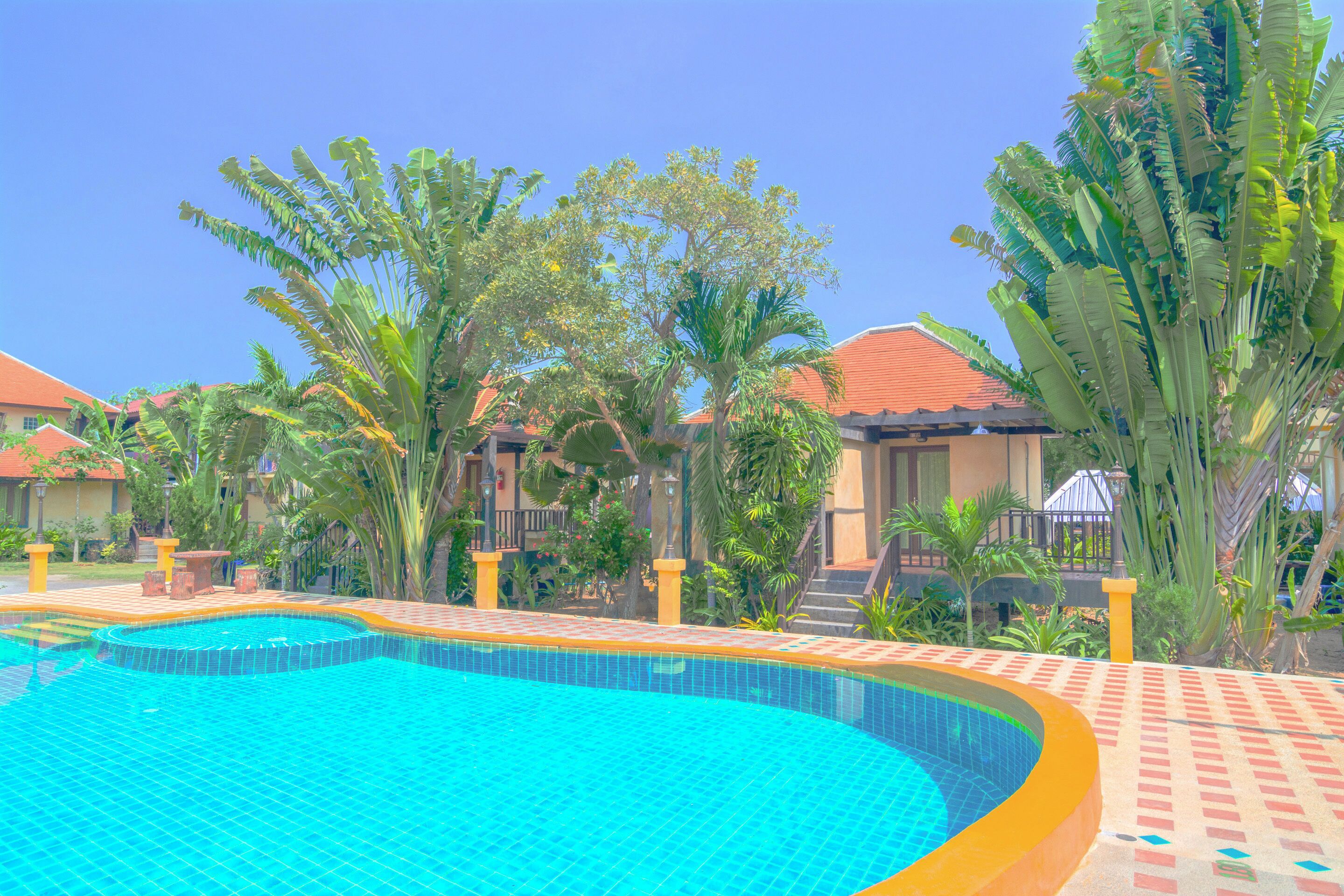 A swimming pool with tiled edges, surrounded by palm trees and a building with a red roof.