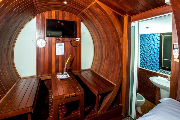 Una habitación compacta con paneles de madera, un banco, una mesa pequeña con un control remoto, un reloj y un baño con pared de azulejos azules.