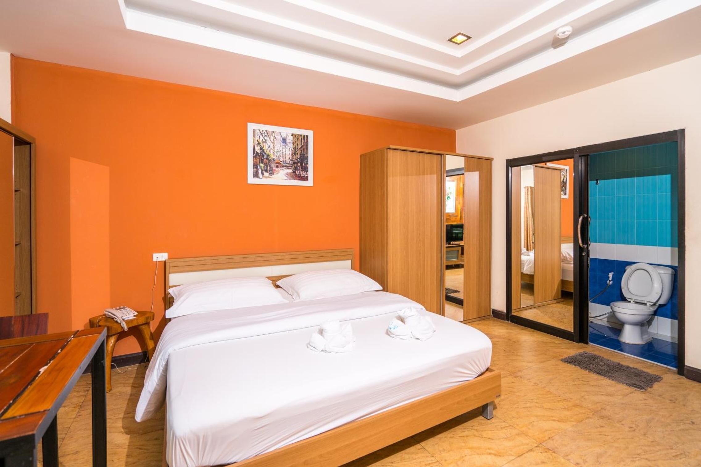 A hotel room with a large bed, a wooden headboard, an orange wall, a small table with a chair, a bedside table, a telephone, and a mirror reflecting a bathroom with a toilet.