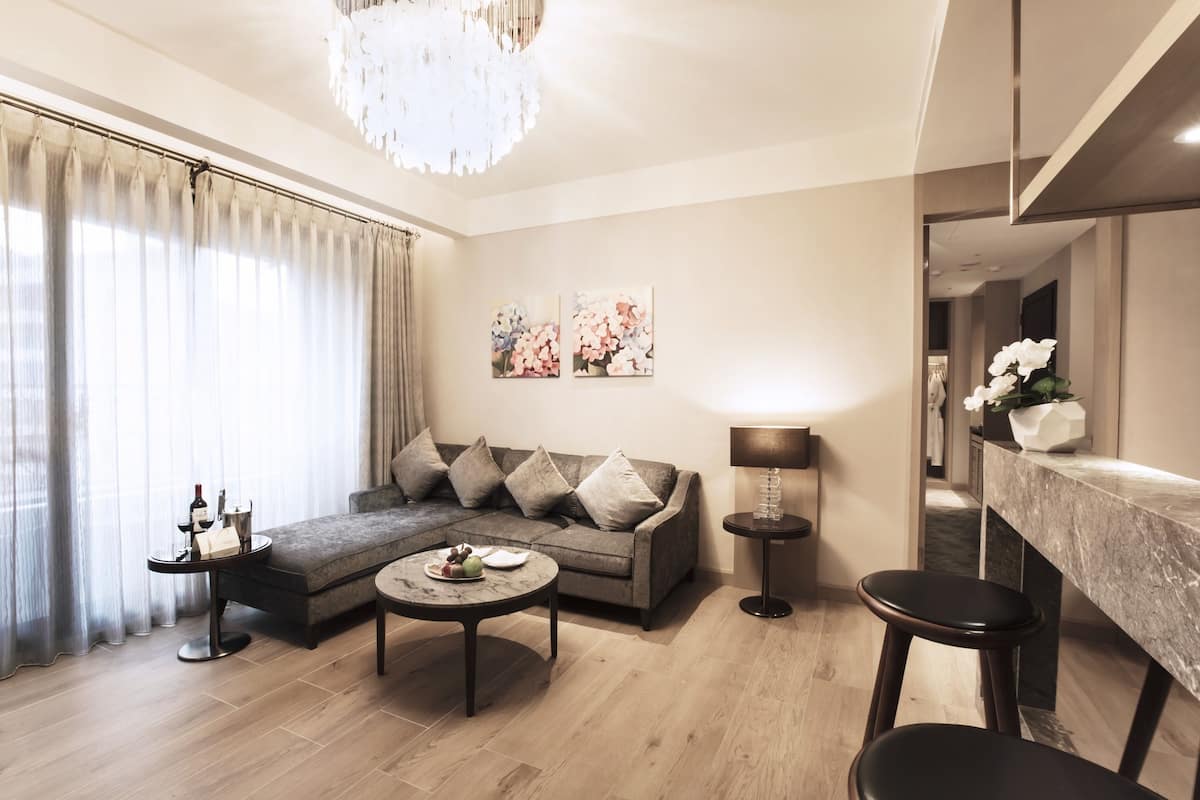 Ein modernes Wohnzimmer mit einem Ecksofa, einem runden Couchtisch, einem kleinen Beistelltisch mit einer Flasche und einer Lampe. Es gibt ein großes Fenster mit Vorhängen, eine Pendelleuchte und zwei gerahmte Blumenbilder an der Wand.