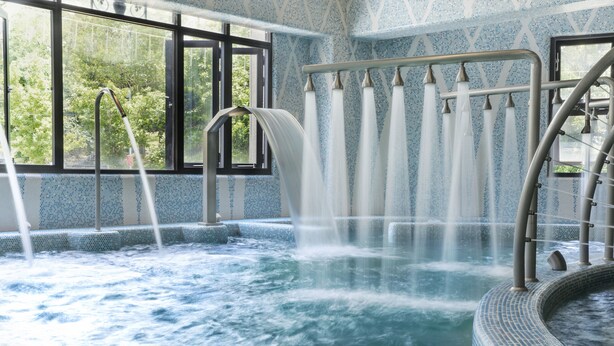 Un jacuzzi con caída de agua, rodeado de baldosas con diseños y ventanas grandes.