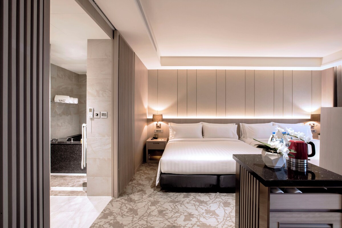 A modern hotel room with a large bed, a bedside table with a red mug, and a nightstand with a lamp.