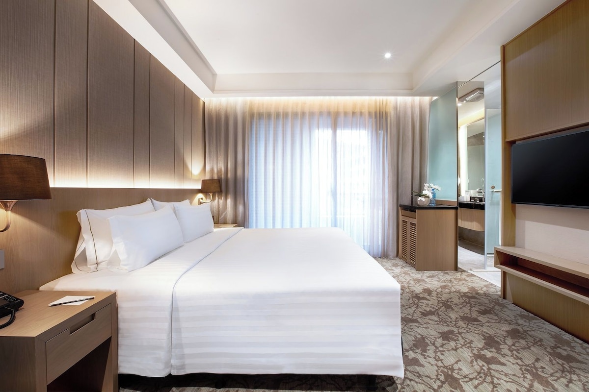 A hotel room with a large bed, a desk with a telephone, a TV, and a bathroom visible through a glass partition.