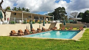 Una piscina con sillones de descanso y sombrillas, rodeada de césped y un edificio con un porche cubierto.