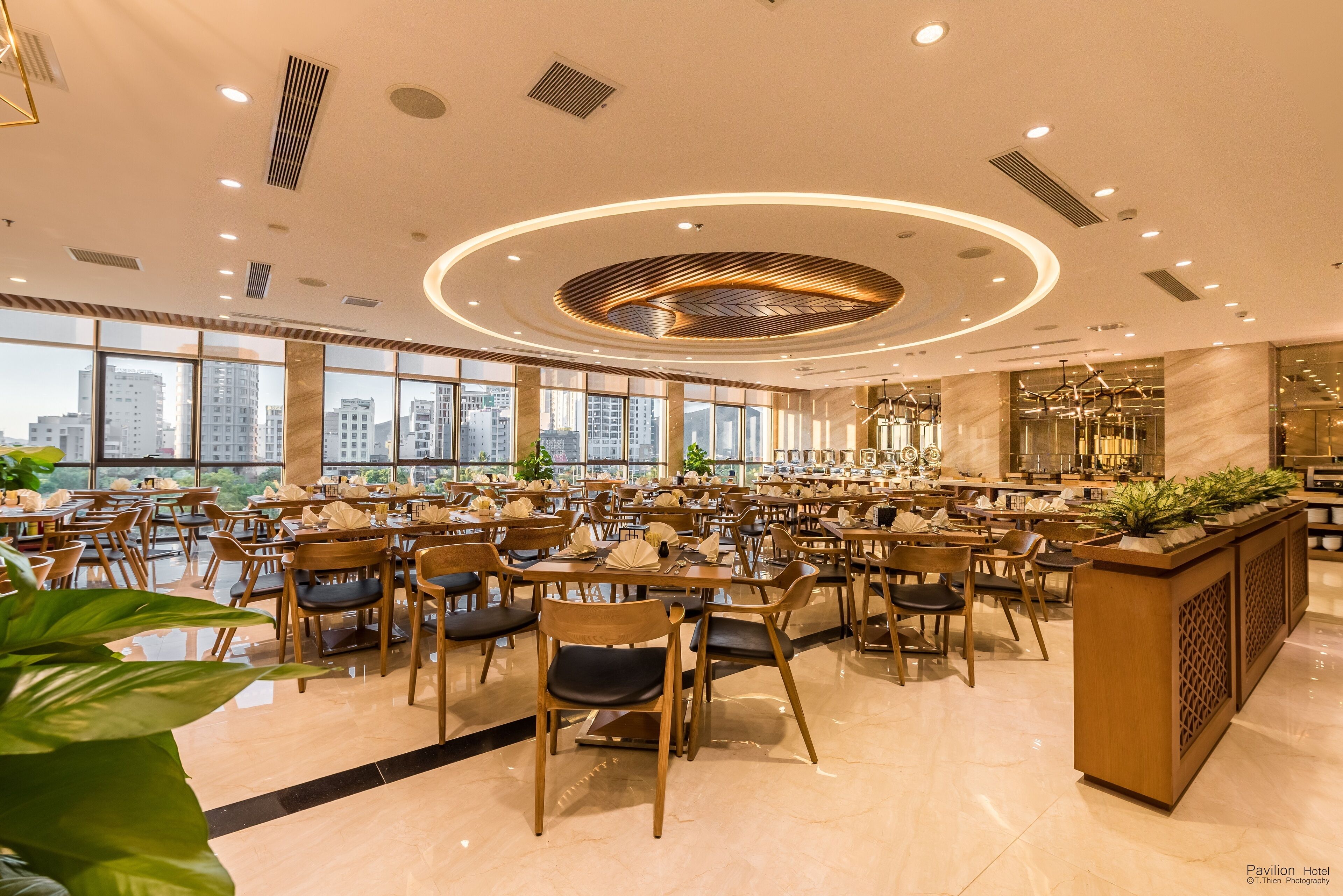 Un'ampia sala ristorante con numerosi tavoli e sedie, ampie finestre che offrono vista sulla città e un elemento decorativo al soffitto.