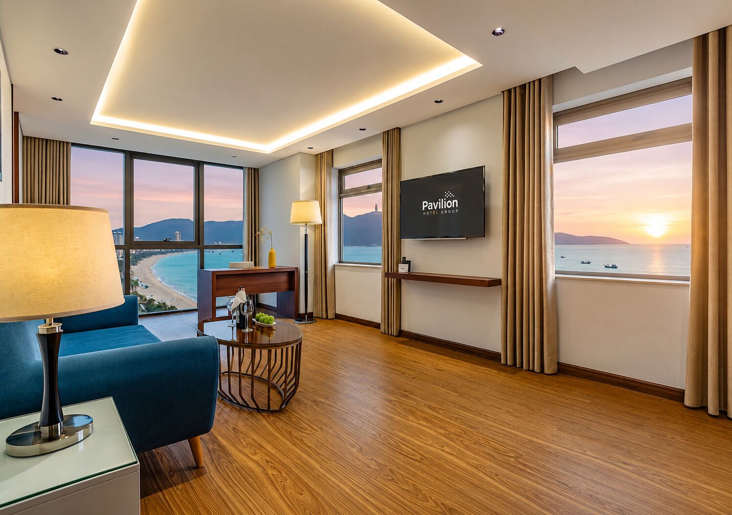 A hotel room with a large window, a flat-screen TV, a blue sofa, a wooden floor, and a table with a lamp.