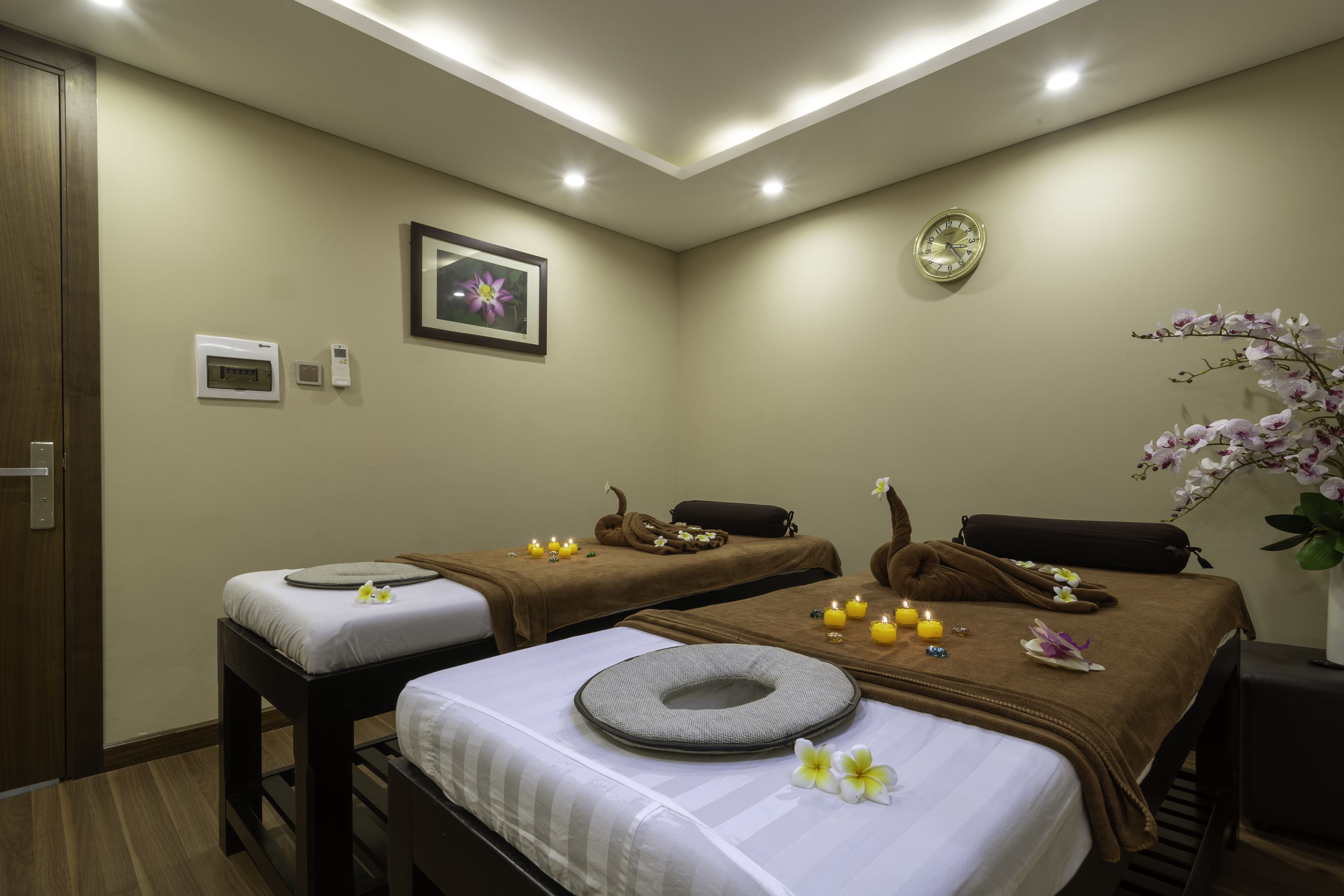 A spa room with two massage tables, each with a towel and a decorative headrest. There are candles and flowers on the tables, and a clock on the wall.