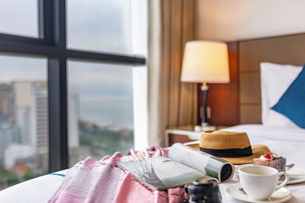A hotel room with a bed, a lamp, a hat, a cup of coffee, and a book on the bedside table.