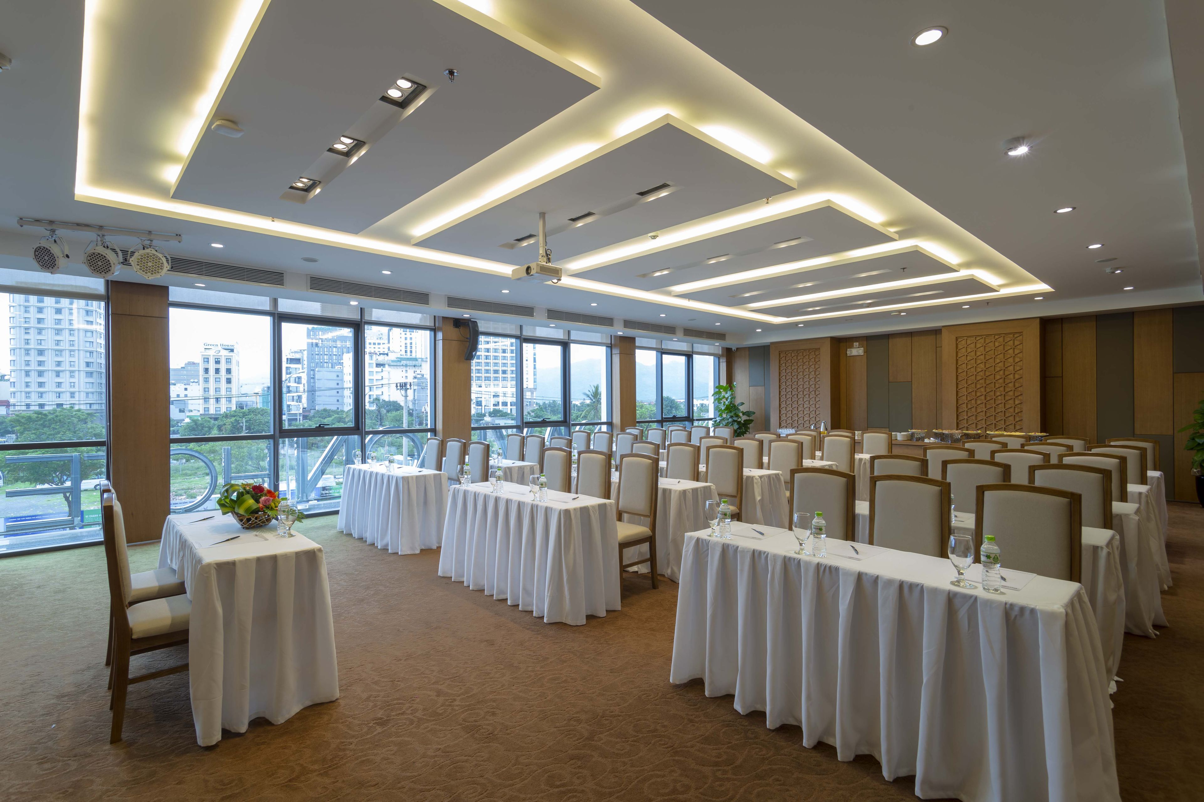 A conference room with multiple tables and chairs, large windows with a city view, and modern lighting fixtures.